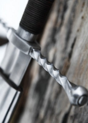 Épée bâtarde à pommeau en coquille cannelée – quillon torsadé et garde acier – gros plan sur la monture contre tronc clair