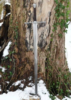 Épée bâtarde à pommeau en coquille cannelée – lame acier longue et garde torsadée – vue complète contre tronc neige et lierre