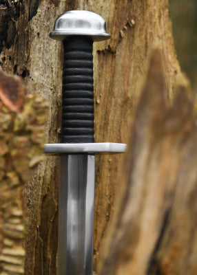 Épée viking à pommeau en bouton – poignée noire, garde et pommeau argentés – vue verticale contre tronc, arrière-plan flou