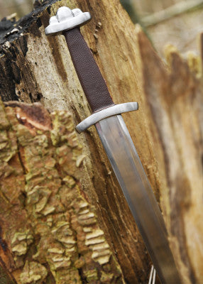 Gros plan sur la garde et la poignée de l’épée viking Wheeler VI SK-C, appuyée contre une souche, rendu médiéval lisible