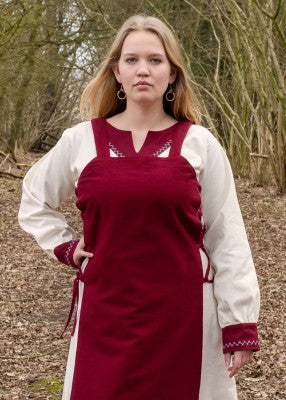 Détail du tablier viking Tinna rouge porté sur une robe blanche, avec encolure droite et bretelles larges